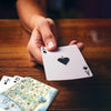 A close-up of a hand holding an Ace of Spades playing card over a wooden table, with other cards and a Oregon map card visible nearby.