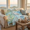 Sofa with a map of Outer Banks, North Carolina blanket draped over it, in a room with large windows overlooking a beach.