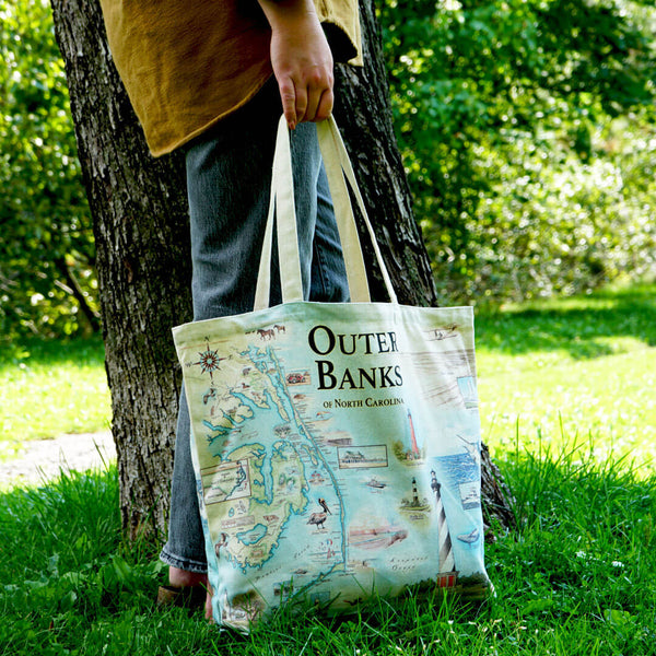 A person holding a Outer Banks canvas tote bag standing in front of a tree.