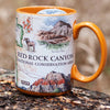 Orange Red Rock Canyon National Conservation Area Ceramic Mug on forest log. Illustrations include Mt. Willson, Rainbow Mountain, Bridge Mountain, Spring Mountain Ranch. Features red-tail hawk, desert Bighorn, Globemallow, Joshua tree, Prickly pear cactus.