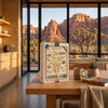 A 1000-piece jigsaw puzzle box titled 'Red Rock Canyon' stands on a wooden table in a modern kitchen setting, with a large window behind it showing a desert canyon landscape at sunset.