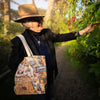 An older woman wearing a tan hat and black jacket stands on a sunlit garden path, holding a San Francisco-themed tote bag and reaching toward red flowers on a plant.