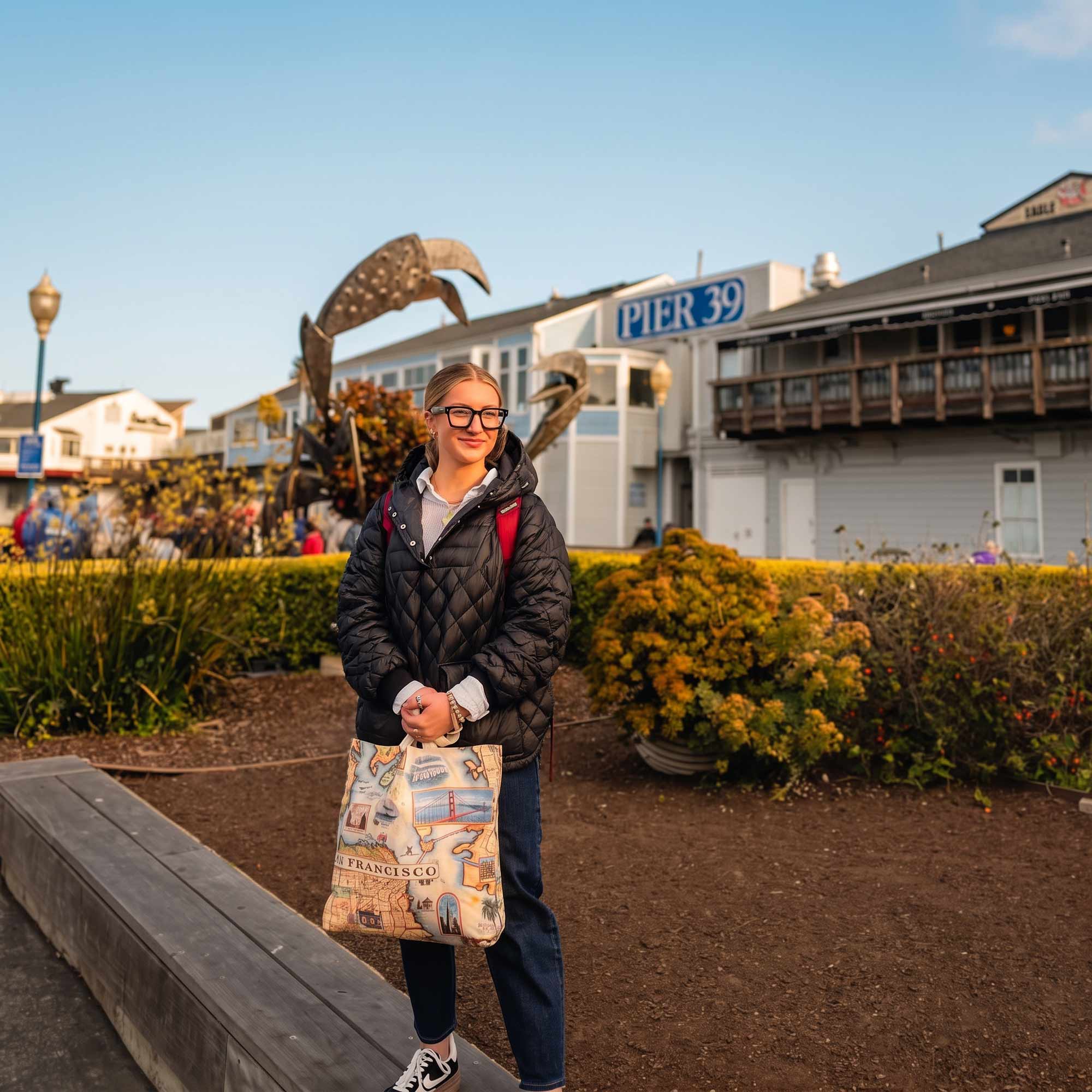 A woman wearing glasses and a black quilted jacket stands outdoors in a park-like setting near Pier 39, holding a canvas tote bag with a vintage-style travel map print.