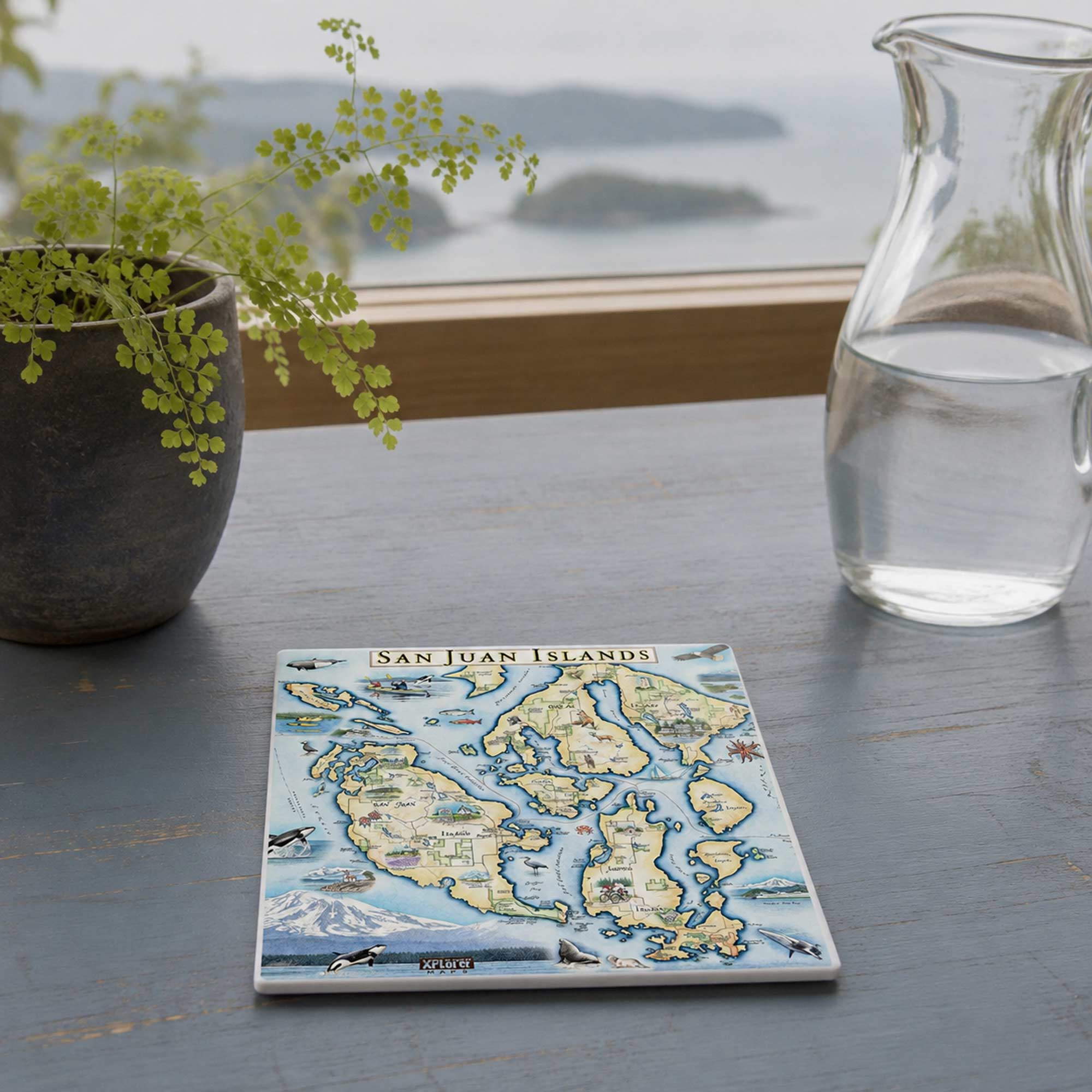 A square ceramic tile coaster featuring a colorful vintage-style map of the San Juan Islands, displayed on a gray wooden table next to a potted fern and a glass water pitcher with a scenic ocean view in the background.