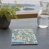 A square ceramic tile coaster featuring a colorful vintage-style map of the San Juan Islands, displayed on a gray wooden table next to a potted fern and a glass water pitcher with a scenic ocean view in the background.