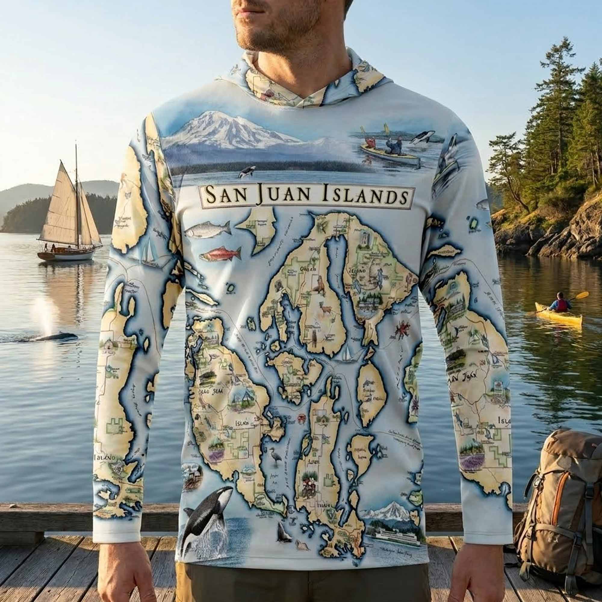 Person wearing a hoodie with a map of the San Juan Islands design, standing by a body of water with a sailboat and kayaker.