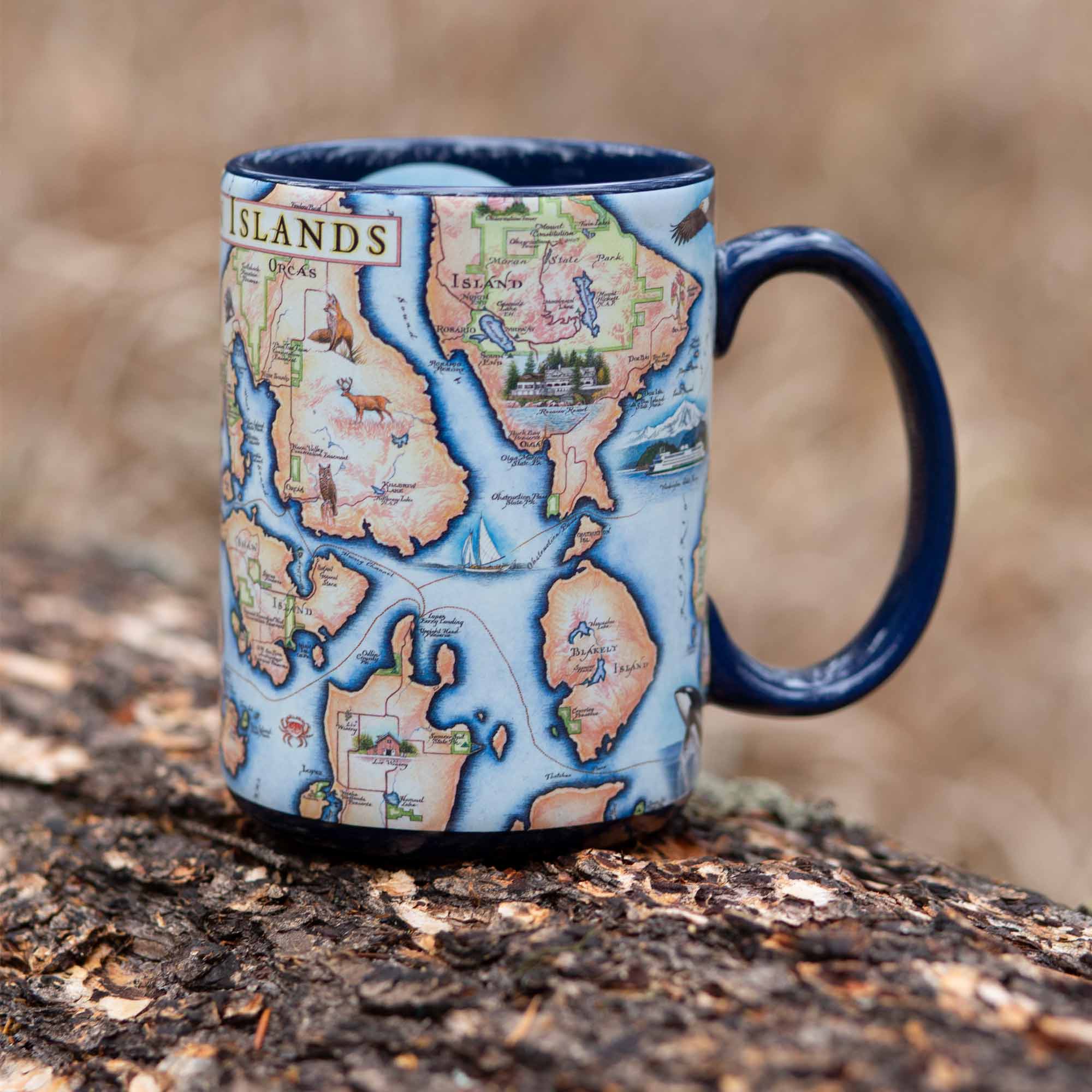 Blue 16 oz San Juan Islands Map Ceramic Mug with handle sitting on a log in the forest. 
