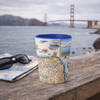 A San Francisco-themed ceramic cup with a vintage map print and blue interior sits on a wooden railing with a folded map and black sunglasses in the foreground, and the Golden Gate Bridge and water in the background.