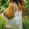 A person outdoors wearing a mustard yellow jacket and carrying a canvas tote bag over their shoulder. The tote bag features a vintage-style illustrated map of the Capiva Sandwich Islands with birds, palm trees, and floral details.