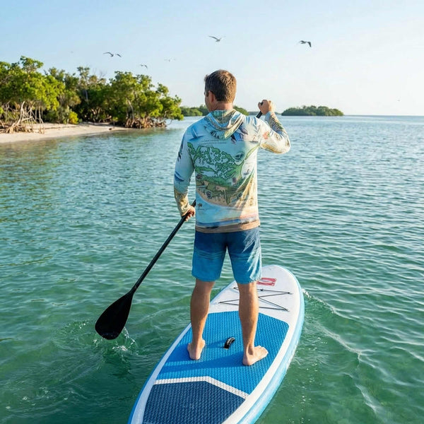 Man paddleboarding on a clear blue water with a tropical island in the background wearing a Sanibel & Captiva Islands sun shirt