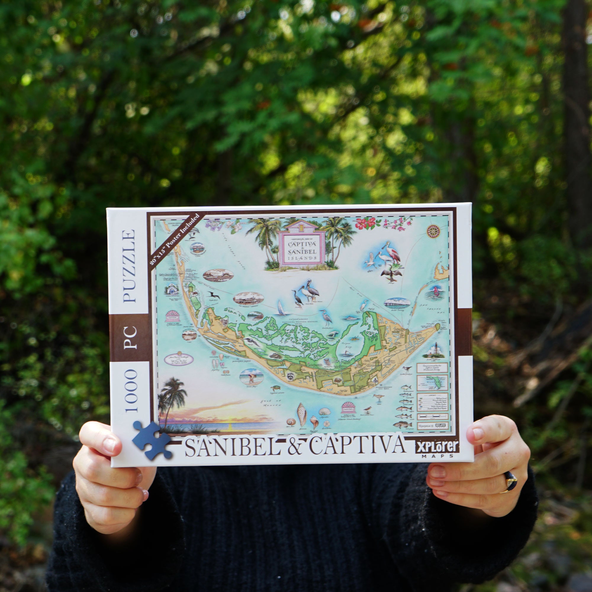 A person holding a completed jigsaw puzzle of Sanibel Island in an outdoor forest setting. The puzzle box is white with a colorful illustration of the island, and the person is wearing a black sweater.