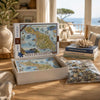A 1000-piece Santa Catalina Island jigsaw puzzle set displayed on a wooden table in a bright, sunlit room. The open white box shows the puzzle map, with the puzzle pieces in a clear bag beside it and matching books stacked nearby.