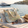A canvas tote bag printed with a vintage-style map of Santa Catalina Island sits on a white wooden beach chair by the water, styled with sunglasses, a rolled towel, and a straw hat.