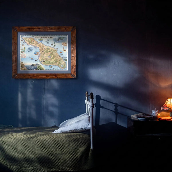 A framed vintage map of Great Gorda Island hanging on a dark blue wall above a bed in a dimly lit bedroom. A bedside table with a lit lamp is visible on the right.