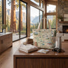 A canvas tote bag printed with a vintage-style map of Kings Canyon National Park stands on a wooden table in a modern lodge setting. The bag is styled with a tan hat, a folded cream shirt, a small notebook, and a stainless steel water bottle, with large windows in the background showing a mountain and tree landscape.