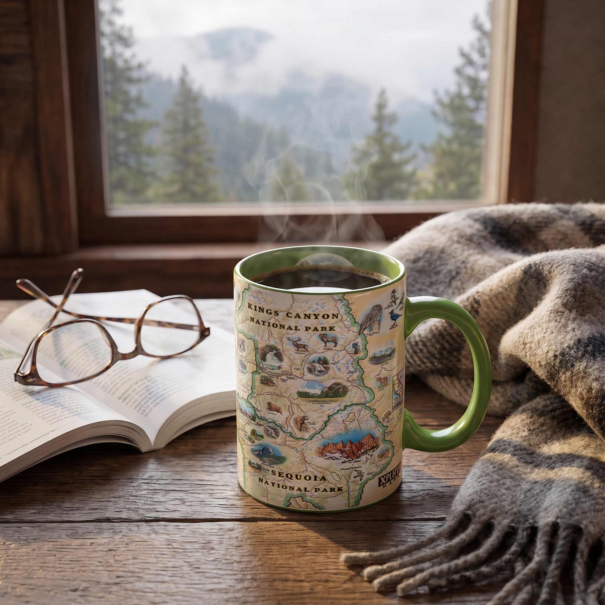 A ceramic coffee mug with a Kings Canyon National Park map design sits on a wooden table next to an open book and glasses, with a cozy gray-and-brown throw blanket and a window showing a misty mountain forest in the background.