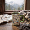 A ceramic coffee mug with a Kings Canyon National Park map design sits on a wooden table next to an open book and glasses, with a cozy gray-and-brown throw blanket and a window showing a misty mountain forest in the background.