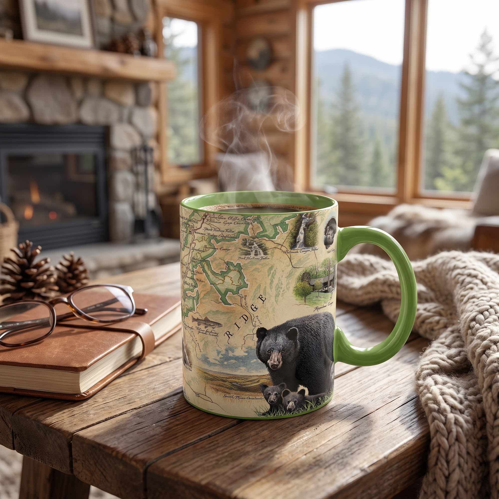 A ceramic mug with a green handle and rim sits on a rustic wooden table in a cozy cabin setting. The mug features a detailed map design with a black bear and cubs at the bottom.