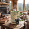 A ceramic mug with a green handle and rim sits on a rustic wooden table in a cozy cabin setting. The mug features a detailed map design with a black bear and cubs at the bottom.