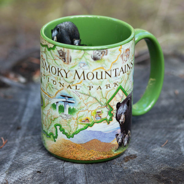 Green 16 oz Great Smoky Mountains National Park Ceramic Coffee Mug sitting on a log. in the forest. The cup Features a black bear, elk, mountain lion, turkey, and Clingmans Dome.