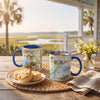 Two ceramic mugs with blue handles and rims are shown on a porch table, one with biscuits and one with steaming coffee, set against a scenic marsh background with palm trees and a vase of yellow flowers.