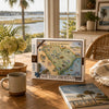 A South Carolina 1000-piece jigsaw puzzle box by Explorers Log Cabin is displayed on a wooden table in a bright, sunny room with a window, a mug, and a matching book in the foreground.