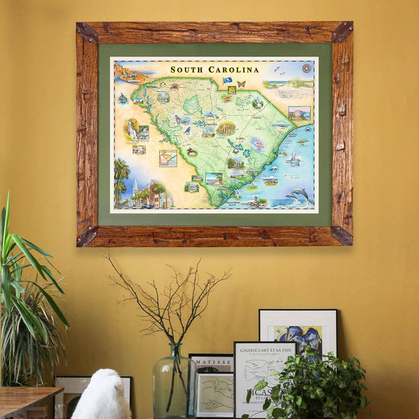 Montana Scraped Pine Frame with Blue Mat featuring a South Carolina map by Chris Robitaille, showcasing landmarks such as Charleston, Myrtle Beach, Hilton Head Island, and Congaree National Park. Columbia, Greenville, and other notable locations are highlighted. The artwork hangs on a mustard yellow wall above framed pictures and plants.