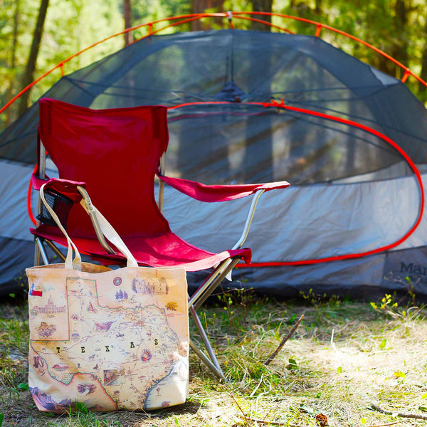 Xplorer Maps' Texas state map canvas tote bag is hanging from a red camping chair outside a camping tent in the woods. 