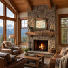 A cozy rustic living room with a large stone fireplace, wood ceiling beams, and mountain views through tall windows. The room includes leather armchairs, a wooden coffee table, a lamp, and framed wall art above the fireplace.