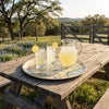 Outdoor setting with a wooden table, pitcher of lemonade, glasses on a Texas servign Tray, and a scenic background.