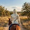Person riding a horse on a dirt path through a scenic landscape with trees and open sky.