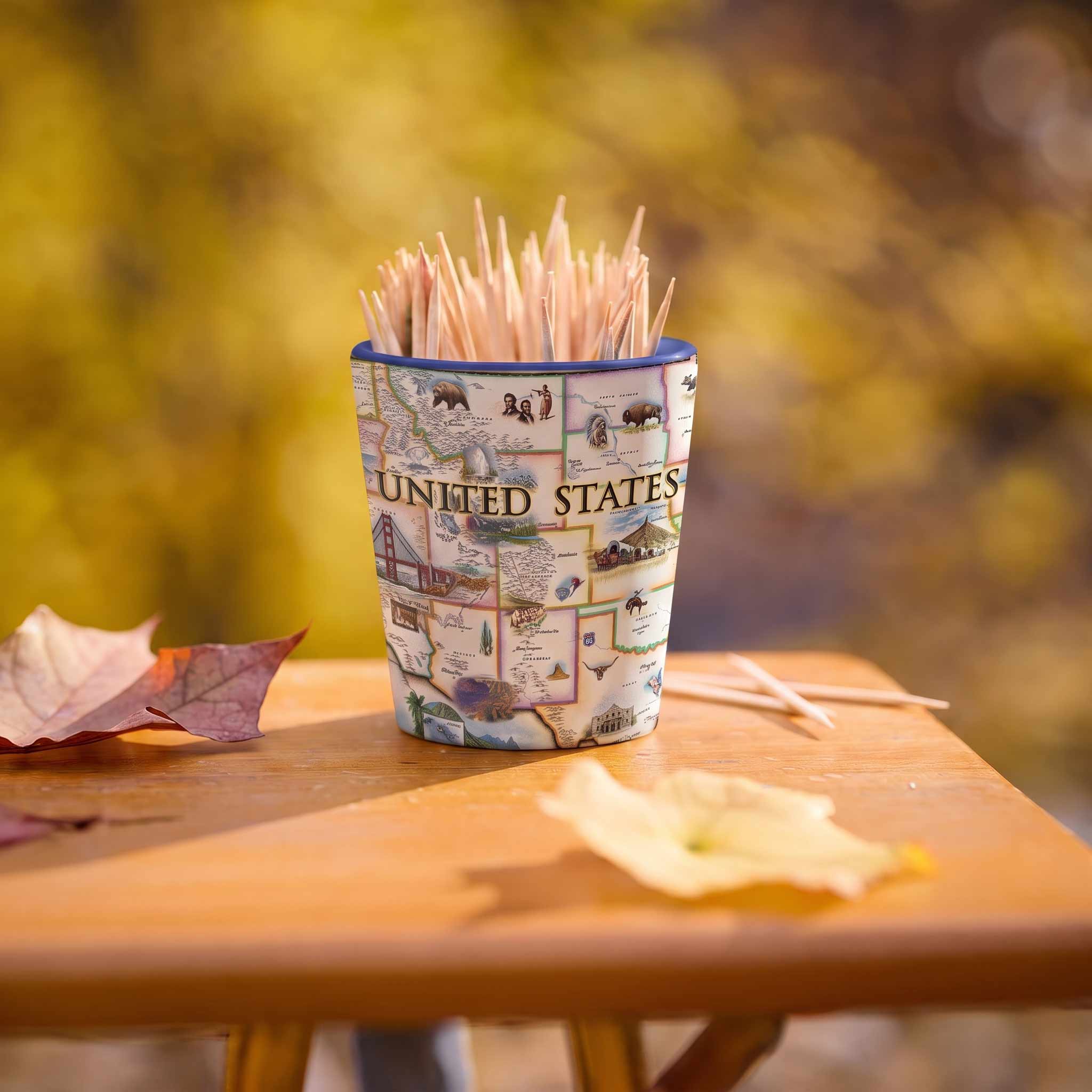 A small cup-shaped toothpick holder printed with a vintage United States map design, filled with wooden toothpicks and placed on a wooden table with autumn leaves and a blurred yellow outdoor background.
