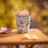 A small cup-shaped toothpick holder printed with a vintage United States map design, filled with wooden toothpicks and placed on a wooden table with autumn leaves and a blurred yellow outdoor background.