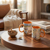 Two ceramic mugs with orange interiors and handles, decorated with scenic collage-style prints, sit on a round wooden table in a warm, sunlit living room. One mug shows the word "UTAH" vertically.