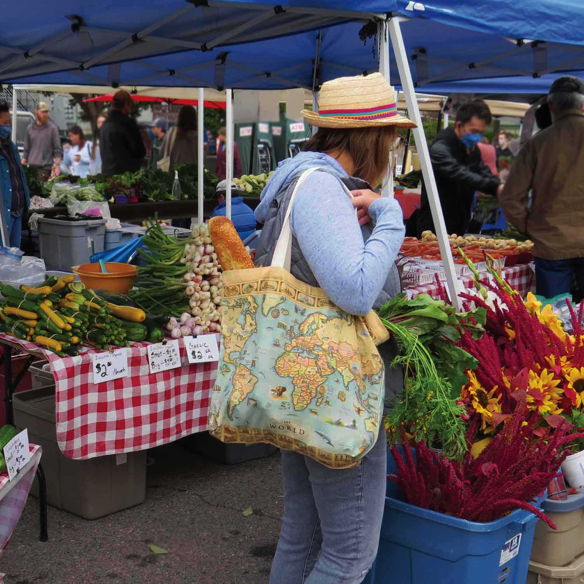 A woman at a farmers market wearing a straw hat and blue hoodie, carrying a large vintage-style tote bag with a world map print. Vegetable and flower stalls are visible under a blue canopy in the background.