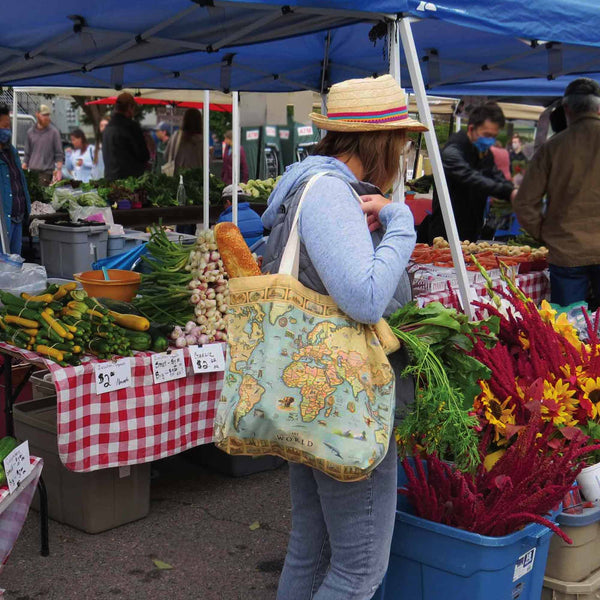 A woman at a farmers market wearing a straw hat and blue hoodie, carrying a large vintage-style tote bag with a world map print. Vegetable and flower stalls are visible under a blue canopy in the background.