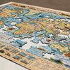 A close-up of a completed wooden world map jigsaw puzzle, showing detailed illustrated continents and oceans with some pieces missing around the edges, photographed on a neutral surface.