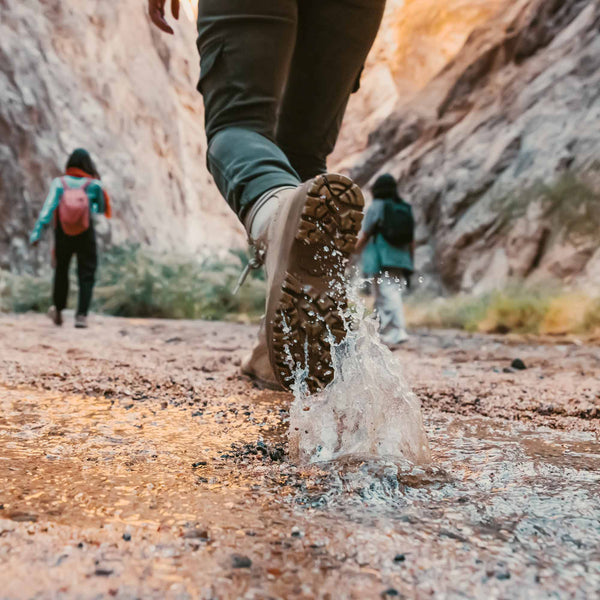 Person is seen stepping through a large puddle, splashing water into the foreground of the image as they go hiking with two other people in a rock canyon.
