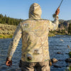 Person wearing a map-patterned sun shirt hoodie fishing by a lake with mountains in the background
