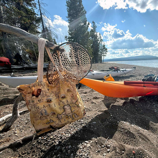 Beige tote bag with map design on a beach with kayaks and fishing net.