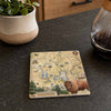 A square ceramic coaster featuring a vintage-style Yellowstone National Park map design with illustrations and a bison in the lower right corner, placed on a dark countertop next to a potted plant and a glass carafe.