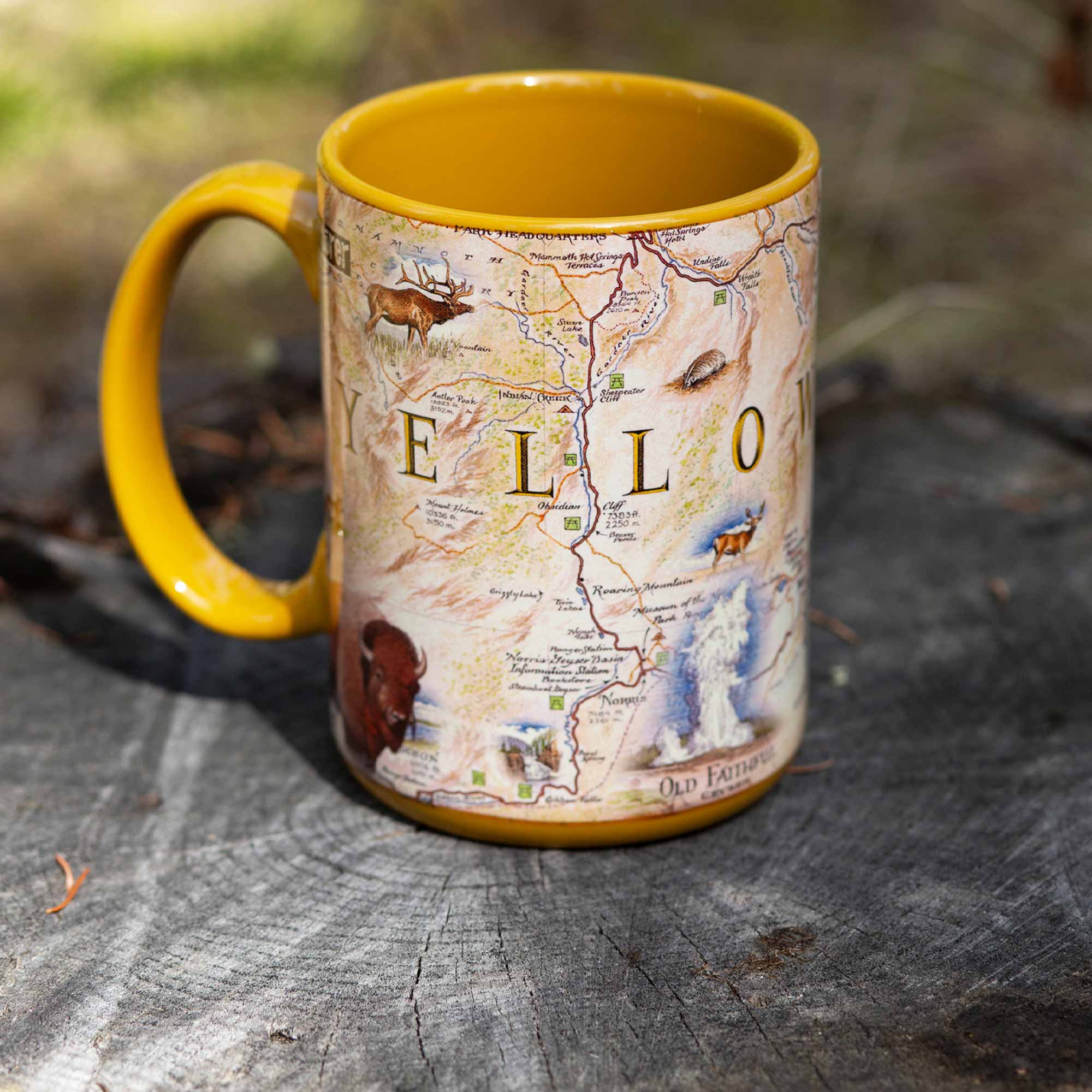 Yellow 16 oz Yellowstone National Park Map Ceramic Mug with handle sitting on a log in a forest.