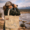 A woman in a brown and beige trucker hat and green plaid shirt looks through binoculars outdoors by a river, carrying a canvas tote bag printed with a vintage map of Yellowstone National Park and an illustration of a bison.