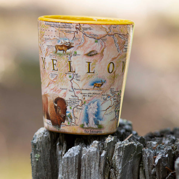 Yellowstone National Park Map Ceramic shot glass by Xplorer Maps sitting on a tree stump. Features illustrations of places such as Yellowstone Lake, Old Faithful, and Roosevelt Tower. Flora and fauna include mountain lions, world, grizzly bears, fireweed, and lupine.