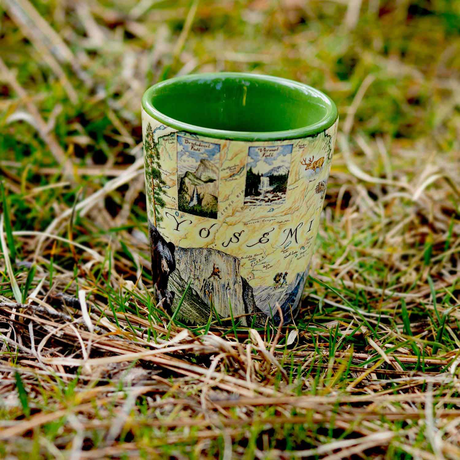 Yosemite National Park Map Ceramic shot glass by Xplorer Maps. The shot is sitting on the grass. Features illustrations of places such as Vernall Falls, El Capitan, and Half Dome. Flora and fauna include mule deer, Indian paintbrush, grey owl, coyote, and a black bear with her cubs. Other illustrations include John Muir, mountain climbing, hiking, and Ansel Adams. 