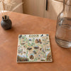 A square ceramic coaster featuring a detailed illustrated map of Zion National Park, styled on a terracotta-colored stone surface next to a glass water bottle and a small vase with dried grass.