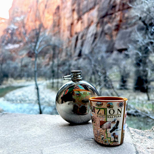 Zion National Park Map Ceramic Shot Glass sitting on a rock ledge in a red rock canyon. Next to the shot glass is a flask. 