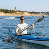 Man paddling a kayak on the water with a lighthouse in the background and wearing a Cape Cod sun shirt hoodie