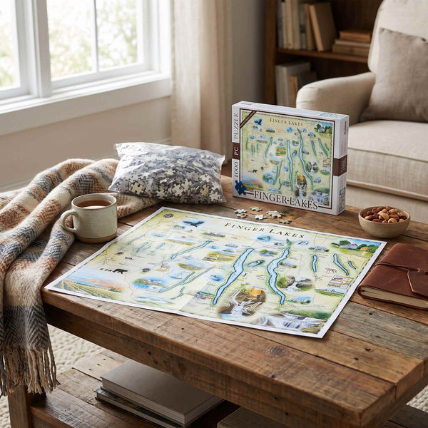 A Fingertips Puzzles 'Finger Lakes' jigsaw puzzle box and completed puzzle are displayed on a rustic wooden table in a cozy living room setting, alongside a mug, blanket, and other home decor.
