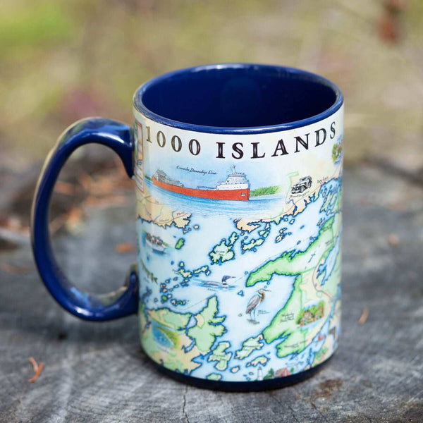 1000 Islands Map Ceramic Mug - 16 oz - Blue, earth tone colors. Map features Bolt Castle and Singer Castle and is sitting on a tree stump.